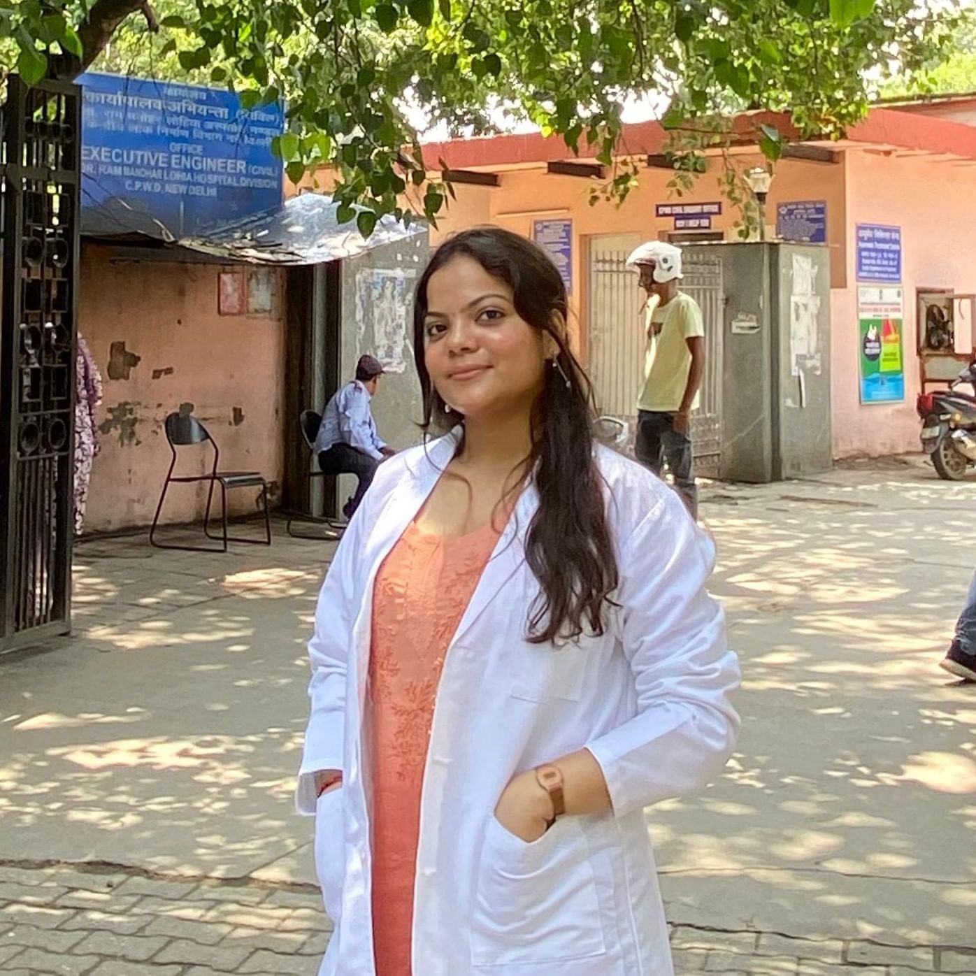 Gitika sharma wearing a white lab coat stands outdoors in front of a building, with hands in pockets. Trees and people are in the background.